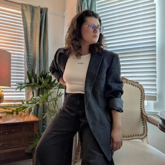 altpfp_mlleone A color photo of Marie Louise "Marlo" Leone, a white woman with dark hair and glasses. She's wearing a dark suit jacket and standing with one hand on her hip, looking over her shoulder.